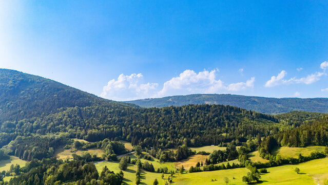 Hillside fields extend between woodland patches and scattered trees.. Green and yellow fields extend below wooded ridges under a blue sky with low white clouds.