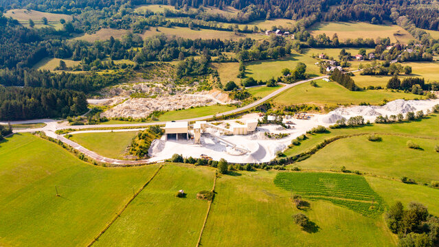 Material stockpiles border fields at a quarry processing site.. Pale buildings at an Austrian quarry border green farmland, with white stockpiles and access roads nearby.