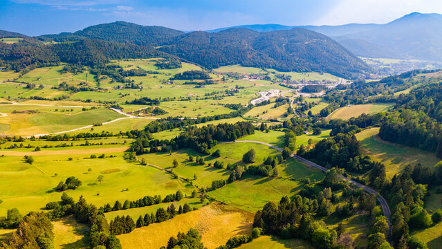 Winding lanes connect scattered houses across valley farmland.. Green and yellow plots follow wooded slopes, with pale roads and roofs viewed from above.