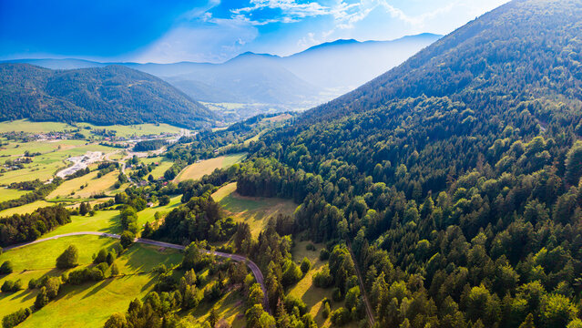 A wooded slope rises above Austrian farmland and village roads.. Green trees fill the right foreground above patchwork farmland, with hazy ridges behind.