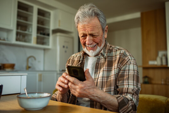 Senior man laughing while texting on smartphone at kitchen table