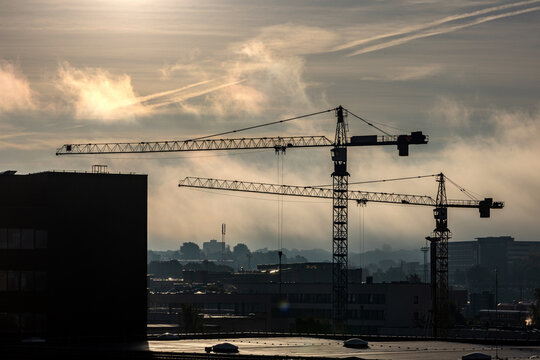 View of construction cranes silhouetted against a misty sky at sunset over the urban rooftops in Vilnius, Vilnius County, Lithuania.