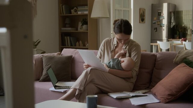 Caucasian young adult mother breastfeeding infant while reviewing business documents. Clip highlights remote work balance, maternity leave, and freelance parenting