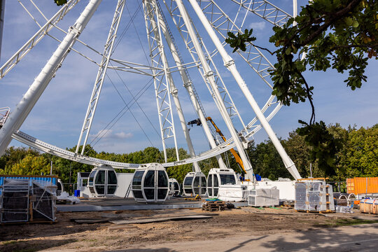 View of a large white Ferris wheel under construction with passenger gondolas, construction equipment, and green trees under a blue sky in Kaunas, Kaunas County, Lithuania.