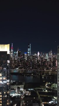 Vertical video: Illuminated New York City skyline captivates at night, with the East River reflecting the vibrant city lights. Skyscrapers and landmarks create stunning urban landscape
