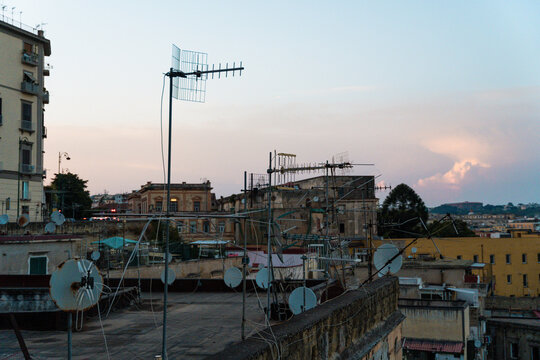 View of a rooftop filled with numerous television antennas and satellite dishes against a soft sunset sky, with historic urban buildings in the background in Naples, Campania, Italy.