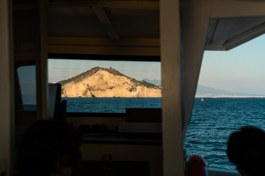 View of a rocky island rising from the sea, framed by the interior of a boat, with calm blue water and clear sky in Procida, Campania, Italy.