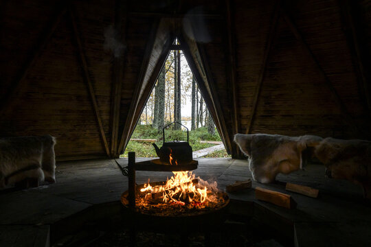 View of a cozy interior of a traditional wooden hut at Apukka Resort with a crackling fire, a kettle, and reindeer hides on benches, overlooking the forest in Rovaniemi, Lappi, Finland.