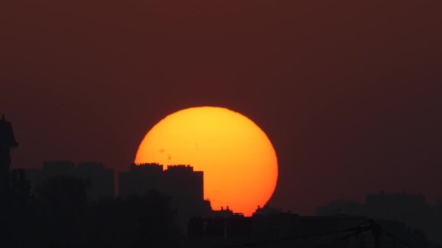 Huge orange sun sets behind a dark city skyline silhouette. Dramatic urban sunset time-lapse with birds flying across the red sky.