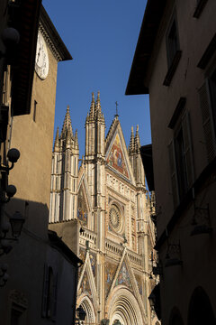 View of the ornate Gothic facade of Orvieto Cathedral with its intricate mosaics and rose window, framed by historic stone buildings in Orvieto, Umbria, Italy.