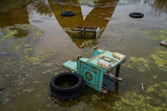 View of a discarded weathered light-blue wooden desk and a black tire submerged in a murky, algae-covered flooded area in Bulgaria.