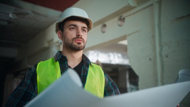 Serious Caucasian contractor man in uniform studying blueprint at construction site male architect builder in helmet holding draft engineer foreman supervising industrial project structure inspection