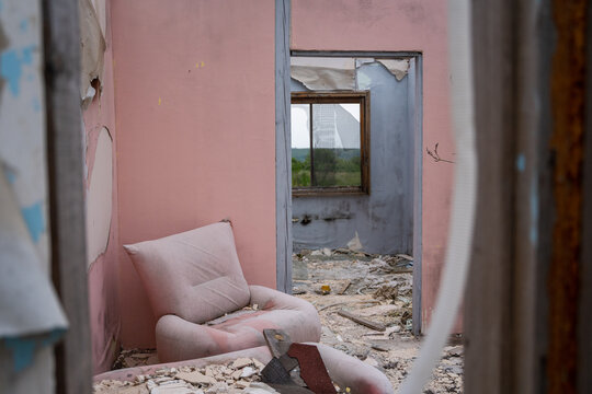 View of a worn pink armchair in an abandoned room with peeling walls and debris on the floor, looking through a doorway toward a broken window in Bulgaria.