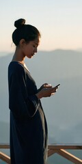 Fototapeta premium Young woman with hair in a bun reading a phone on a mountain balcony while holding a glass — serene outdoor profile portrait background for wallpaper and banner