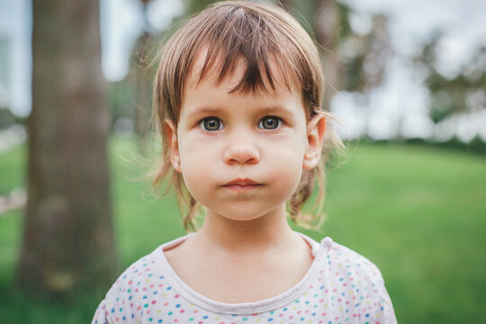 Close up portrait of serious toddler girl with big gray green eyes, brown hair, bangs in polka dot top looking at camera in green park, concept of child psychology, emotions, pediatric health