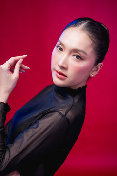 Confident Asian woman looking at camera with striking beauty makeup, showing flawless skin, glossy lips, elegant gesture, and modern glamour charm in dramatic luxury studio lighting portrait
