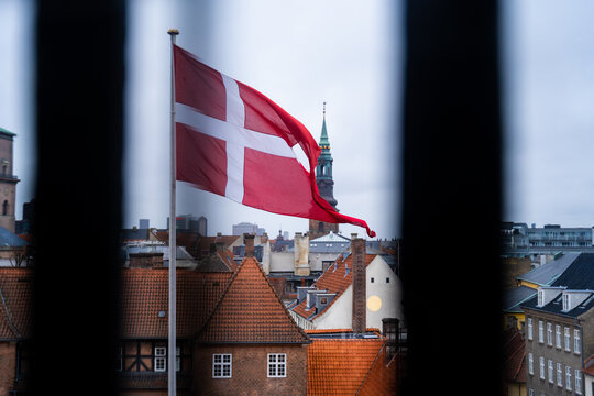 View of the Danish flag waving in the wind against a backdrop of historic rooftops and a church spire in Copenhagen, Denmark.