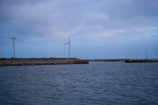 View of wind turbines standing along the coastline near the harbor with calm water in the foreground under a cloudy sky in Copenhagen, Denmark.