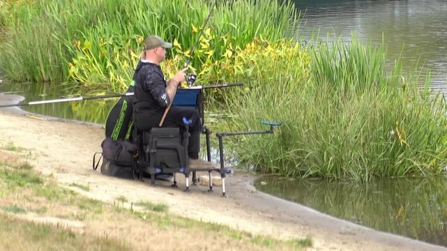 Angler sitting on fishing platform with feeder rods fishing on river bank with green reeds in autumn. Carp fishing and outdoor hobby concept. Fisherman with tackle on sandy shore of calm water.
