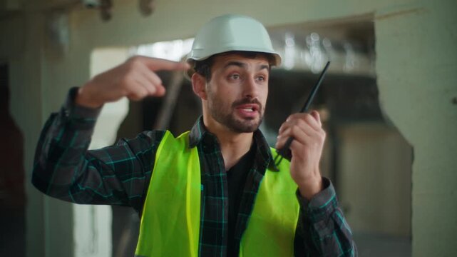 Strict Caucasian builder man pointing gesturing during radio communication on construction site male electrician talking walkie talkie checking engineer supervising foreman instructing technician task