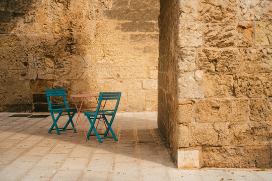 View of a small pink table and two teal chairs set on a stone-paved patio beside a weathered historic stone wall in Bari, Apulia, Italy.