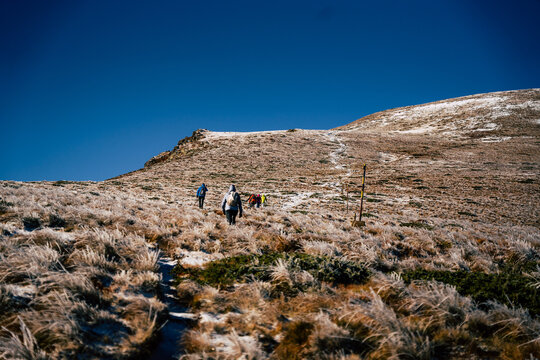 View of hikers trekking up the snowy mountain trail towards the Golyam Kademliya Shelter, surrounded by dry grass and rocky terrain under a clear blue sky, Stara Zagora, Bulgaria.