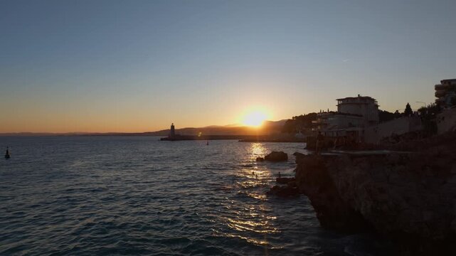 Static sunset behind hills over the Mediterranean, port lighthouse on the jetty and a cliff jumper splashing into the sea, Nice, French Riviera, Cote d Azur, France