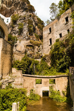 View of historic stone buildings nestled in a rocky gorge with lush greenery and a small waterfall in Atrani, Campania, Italy.