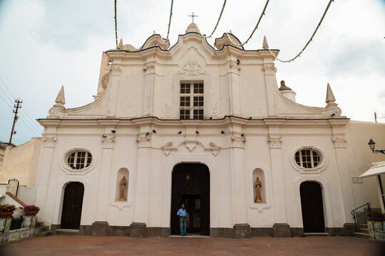 View of the white facade of the Chiesa di San Michele Arcangelo with a man standing in the entrance, decorated with statues and arched windows in Capri, Campania, Italy.
