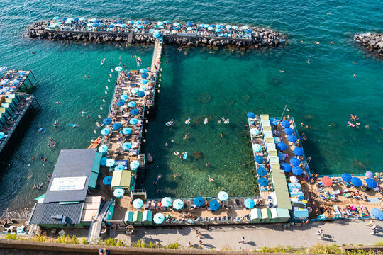 View of a beach club with wooden piers, rows of blue umbrellas, and people swimming in the clear turquoise water in Salerno, Campania, Italy.