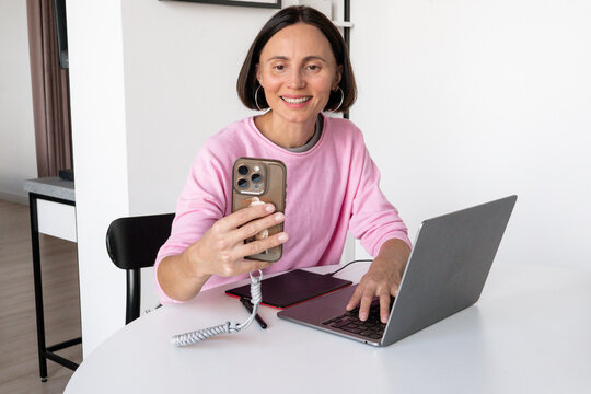 Happy millennial woman in glasses smiling during video call on smartphone, cheerful female digital creator sitting in casual pose at home office desk stock photo