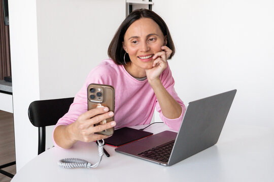 Happy millennial woman in glasses smiling during video call on smartphone, cheerful female digital creator sitting in casual pose at home office desk stock photo