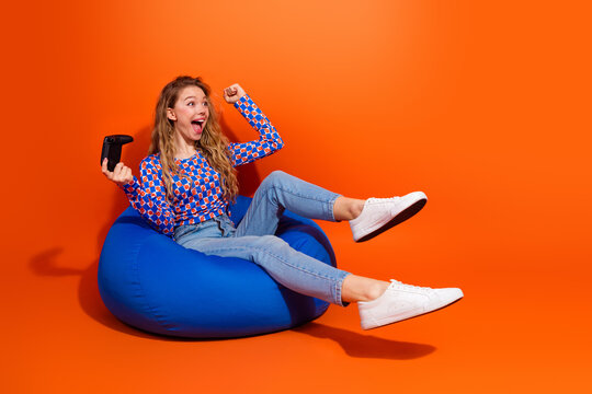 Excited young woman celebrating with a gaming controller while sitting on a beanbag against an orange backdrop
