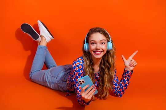 Cheerful young woman with headphones relaxing with a smartphone against a vibrant orange backdrop