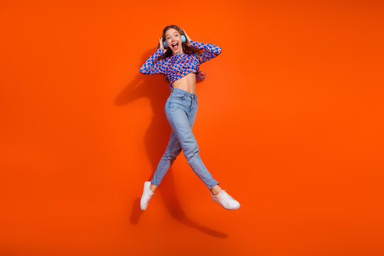 Charming woman enjoying music with headphones, jumping in excitement against an orange backdrop, trendy casual outfit