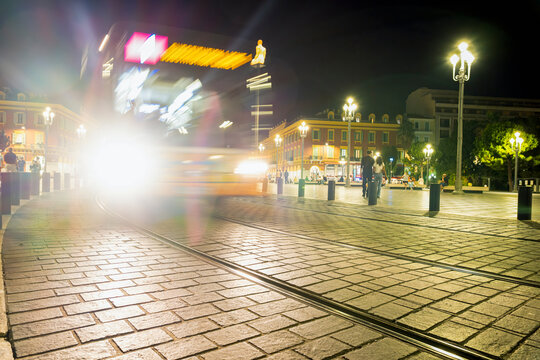 City bus moving through Place Massena in Nice, France, captured from a low angle at dusk with motion blur and light trails, illustrating urban public transport and city traffic
