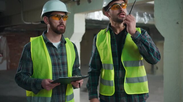 Two serious Caucasian men discussing industrial project at factory males foreman using walkie talkie architect in uniform coordinating mechanics builders engineers supervising construction teamwork