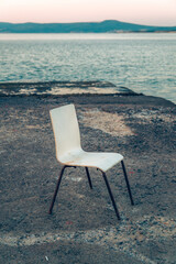 Fototapeta premium A single white chair standing alone on a concrete seaside pier at dusk with calm sea waters in the background.