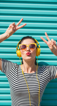 Woman wearing sunglasses and headphones, pouting lips, holding up peace signs against a blue striped background, conveying fun and positivity