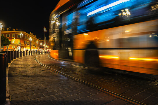 City bus moving through Place Massena in Nice, France, captured from a low angle at dusk with motion blur and light trails, illustrating urban public transport and city traffic