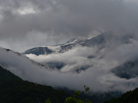 Snowy peak revealed through parting storm clouds, sunlit ridge and dark forest below