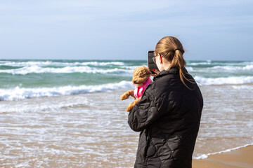 A young woman stands on a sandy beach holding her small dog and recording the beautiful sea waves on a cellphone. Cozy autumn or winter seaside travel landscape. Travel theme. © Natalia