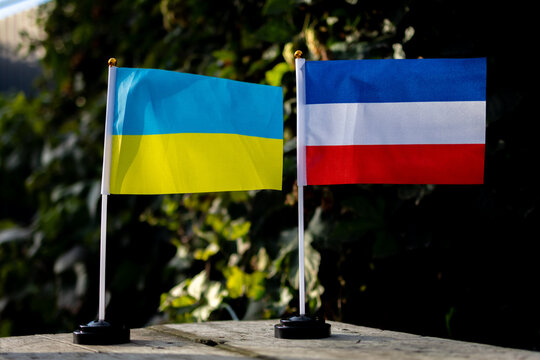 Ukraine and Crimea miniature flags on desk poles rise above a weathered board with leafy bokeh and bright sun creating a balanced scene for neutral reporting education travel and cultural discussion