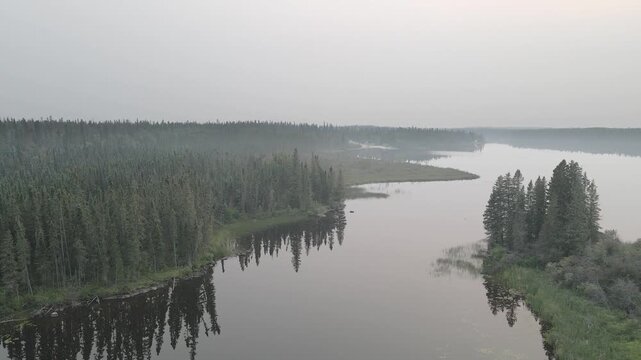 Drone moving above a calm lake covered with forest fire smoke and surrounded by evergreen trees
