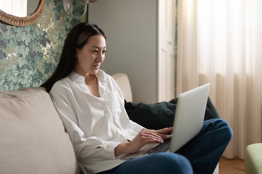 Asian woman using laptop on sofa while resting on couch at home