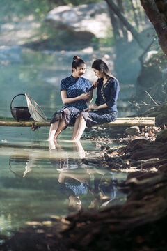 Side view of Two smiling young Thai women wearing traditional indigo blue dresses and patterned sinh skirts sitting on a bamboo bridge with a fishing net fishing, Sakon Nakhon, Isan region, Thailand