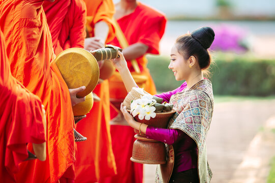 Side view of a smiling Young Laotian woman in a traditional silk dress giving sticky rice morning alms (Tak Bat) to Buddhist monks, Laos