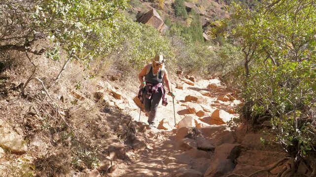 Active senior woman carefully navigating steep, rocky path during a challenging hike in a sunny canyon, using trekking poles for stability and support on outdoor journey. Climbing on top of pass