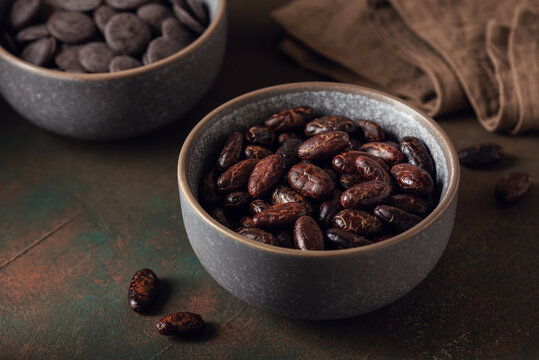 Shelled cacao beans and chocolate buttons in a bowl on a dark rustic background, selective focus.