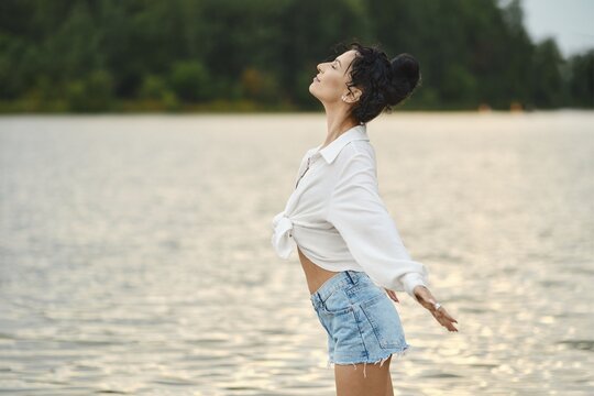 Woman stands near edge of lake with arms open wide as she looks up. She wears white shirt and denim shorts. The sun sets behind her, casting warm light on the water and surrounding trees